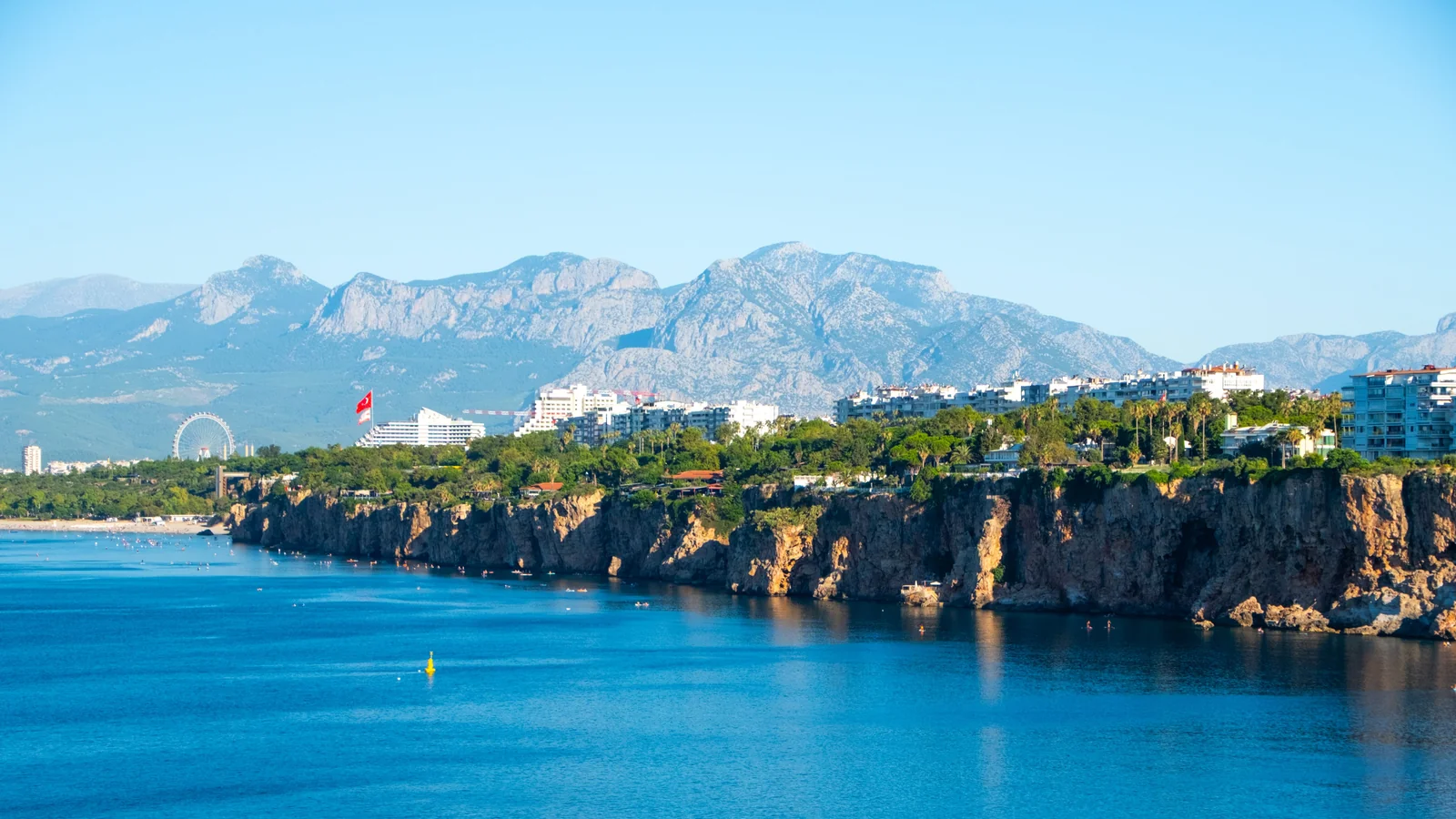 Antalya’s western coastal cliffs with the city, an observation wheel and mountains beyond the blue Mediterranean.