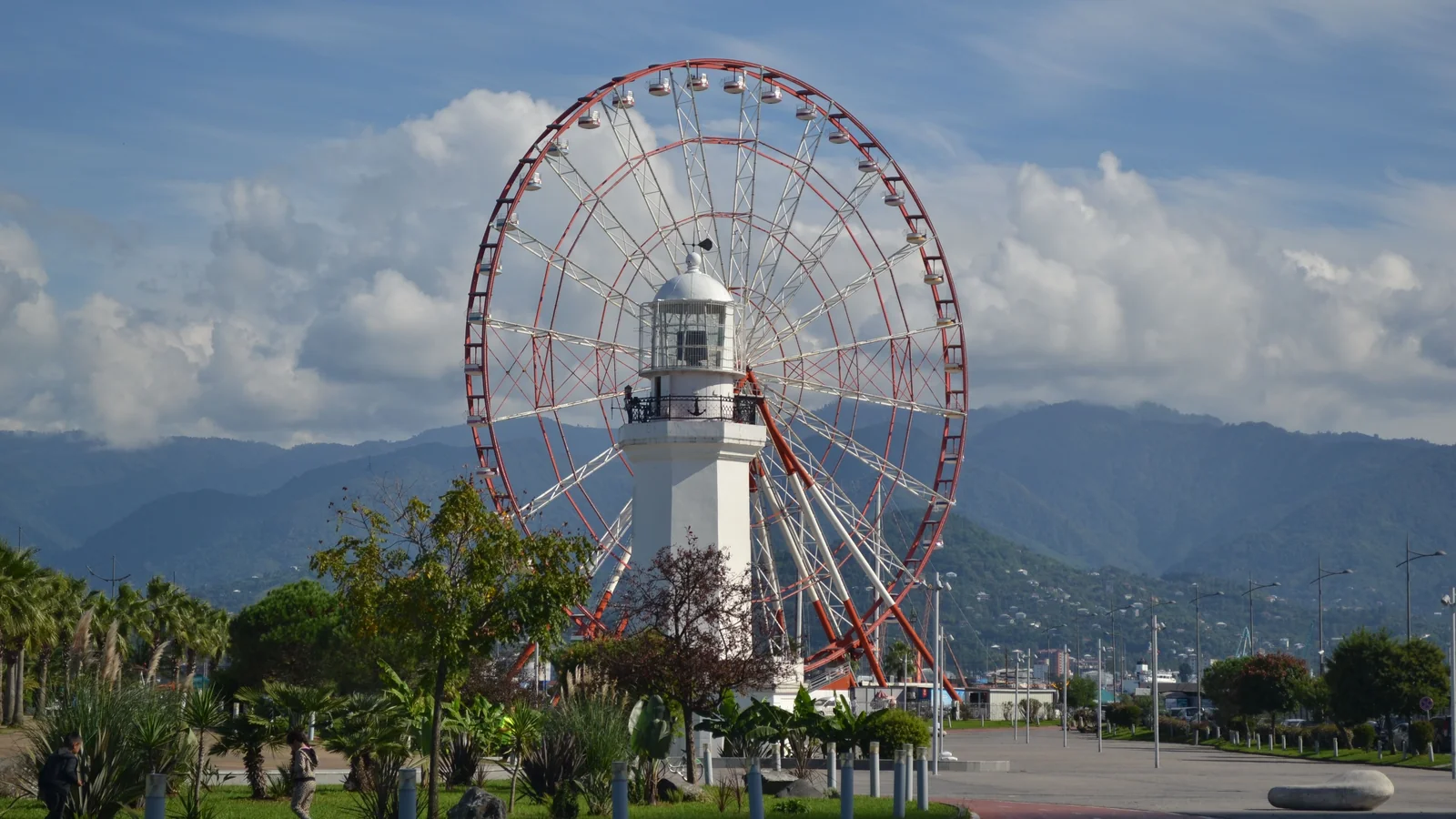 Batumi Lighthouse in front of the Ferris wheel at Miracle Park, with mountains beyond.