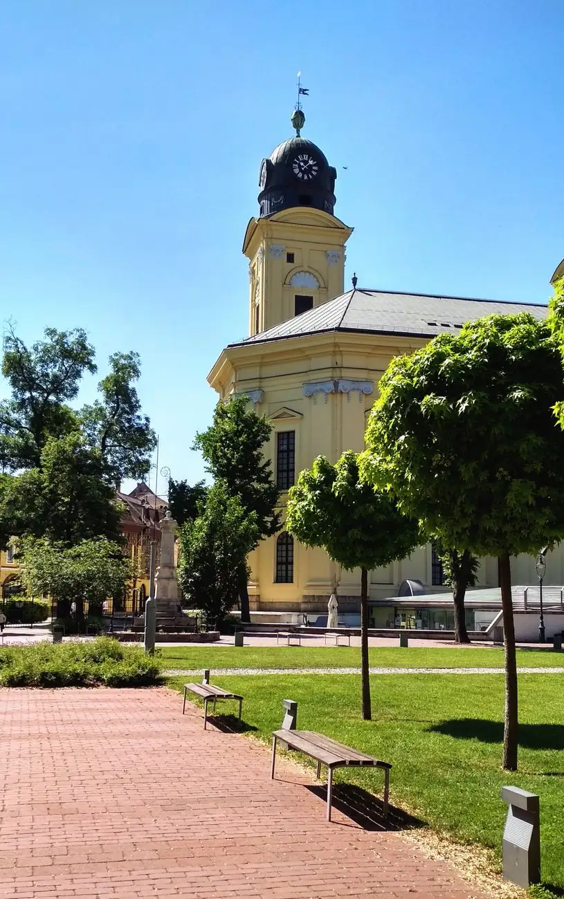 A yellow tower and side façade of Debrecen’s Reformed Great Church beside trees and a paved path