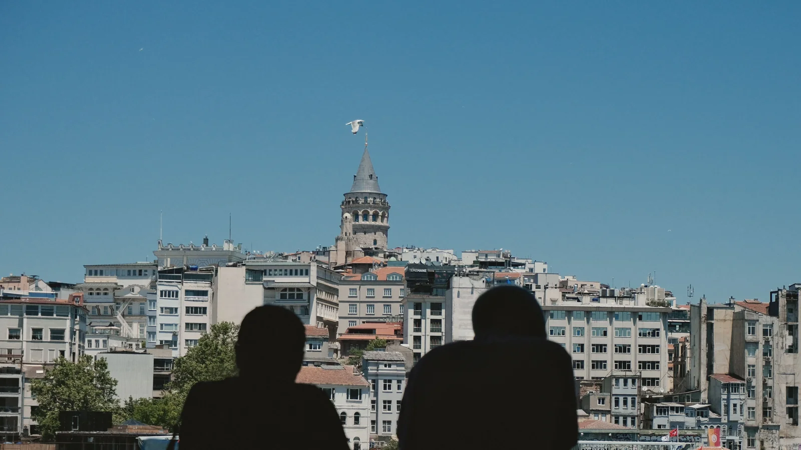 Galata Tower above the Karaköy waterfront, with two silhouetted viewers in the foreground.