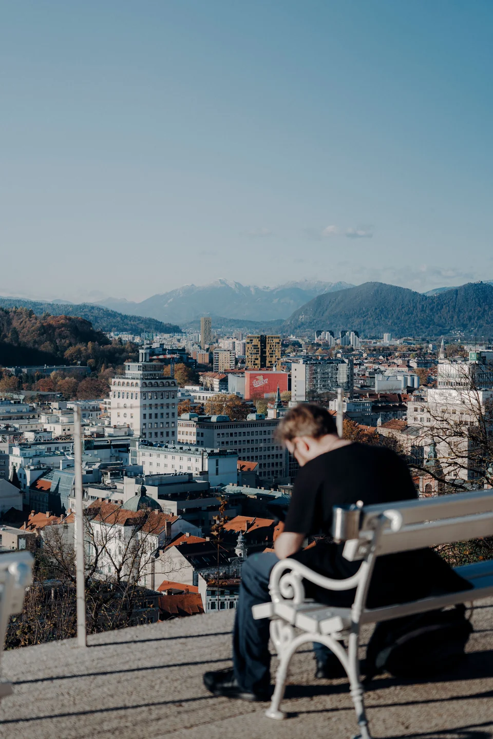 A visitor on a Castle Hill bench overlooking Ljubljana, with Nebotičnik visible in the skyline