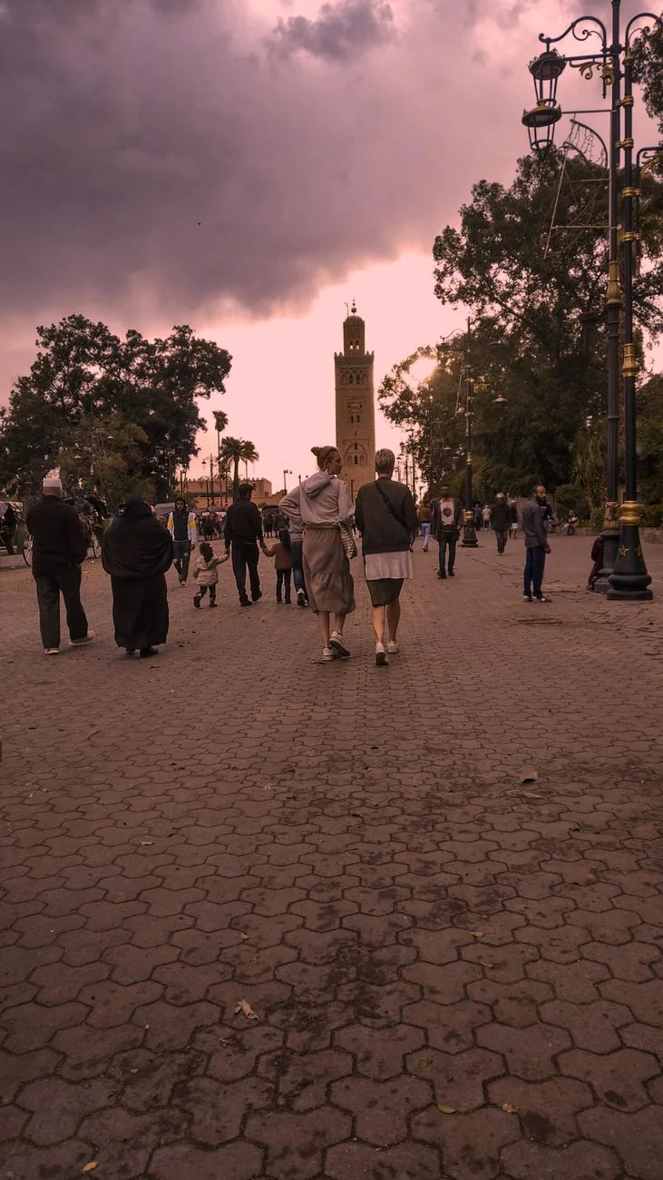 Busy street scene in Marrakech with Koutoubia Mosque in view and people walking under a dramatic sky.