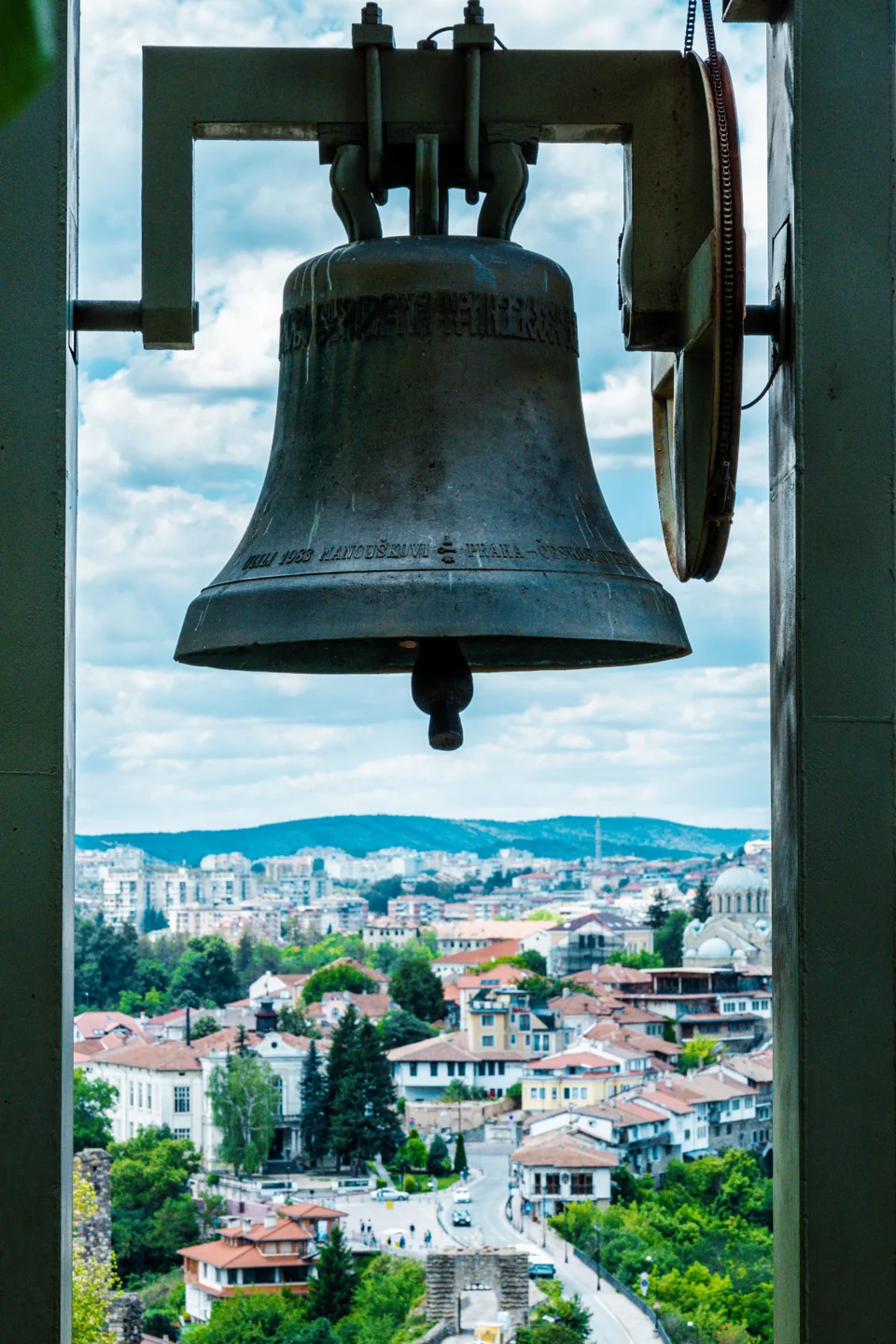 A bell above the Tsarevets entrance approach framing Veliko Tarnovo’s hillside buildings