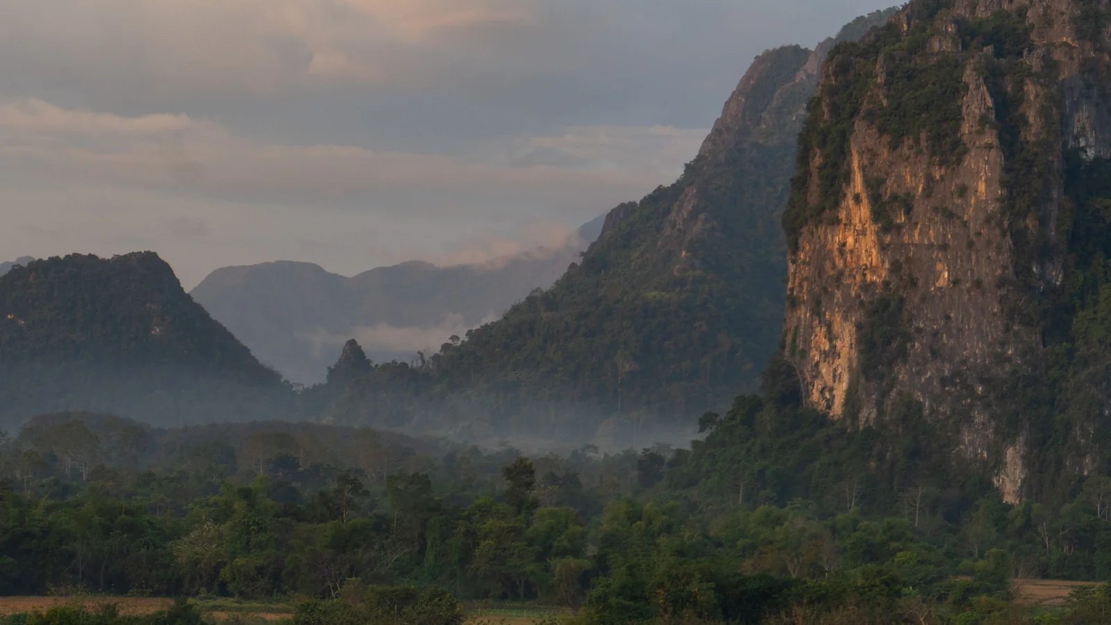 Warm light on Vang Vieng’s limestone cliffs, with forest and mist between the distant hills.